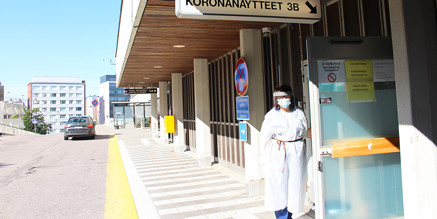 Mia Aho står i skyddsutrustning vid sjukhusdörren. Skylten lyder: Corona prov.